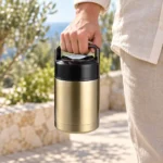 Main portant le pot isotherme inox doré par sa poignée rabattable, sur une terrasse au bord de la mer